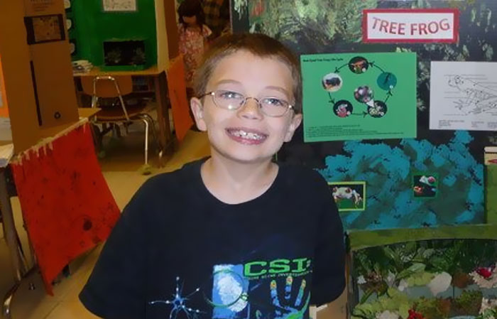 Young boy smiling in a classroom near a science project about tree frogs, relating to unsettling mysteries and fascination.