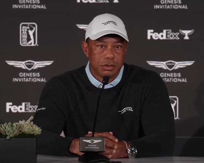 Tiger Woods speaking at a press conference during Genesis Invitational, wearing a white cap and black sweater.