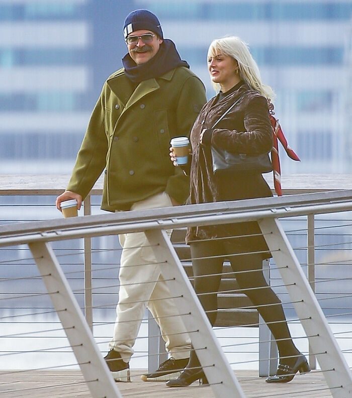David Harbour’s alleged partner walking outdoors holding coffee cups beside a railing with city buildings in the background.