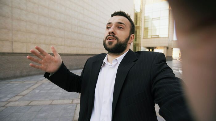 Man in a black suit and white shirt making a confident comeback gesture outdoors on a city street.