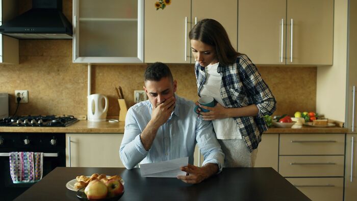 A man looking stressed reading a letter in a kitchen while a woman leans over holding a mug beside him.