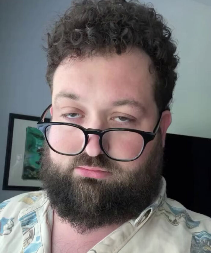 Man with curly hair and beard wearing glasses with a Gen Z stare expression in an indoor setting.