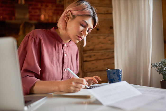 Young woman with pink hair writing notes and researching mansplaining on laptop in cozy wooden room with coffee mug.