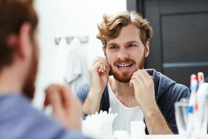Man flossing teeth in bathroom mirror, illustrating common mansplaining moments where men shoot themselves in foot.