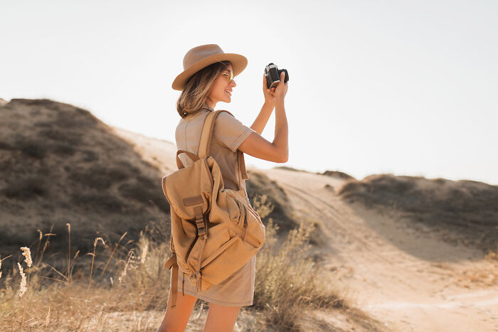 Woman wearing a hat and tan dress, holding a camera outdoors in a dry landscape, capturing moments of mansplaining stories.