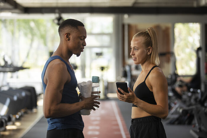 Man and woman talking in gym, woman holding phone as man mansplains during a conversation about fitness and workouts.