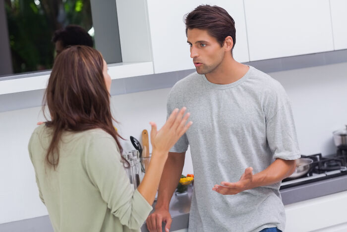 A man mansplaining to a woman in a kitchen, gesturing while she listens with a frustrated expression.