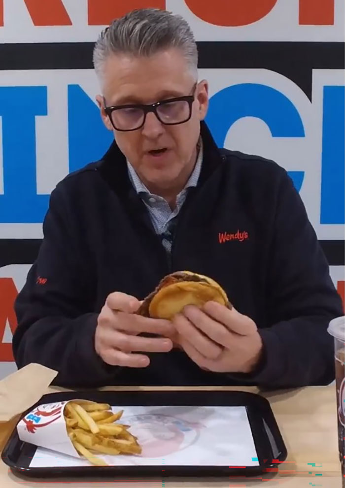 Man wearing glasses and Wendy's jacket holding a burger with fries and drink on tray, highlighting Wendy's burger battle.