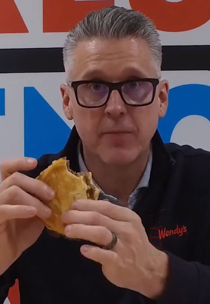 Man wearing glasses and Wendy's jacket holding a burger, symbolizing Wendy's joining the burger battle after McDonald's CEO incident.