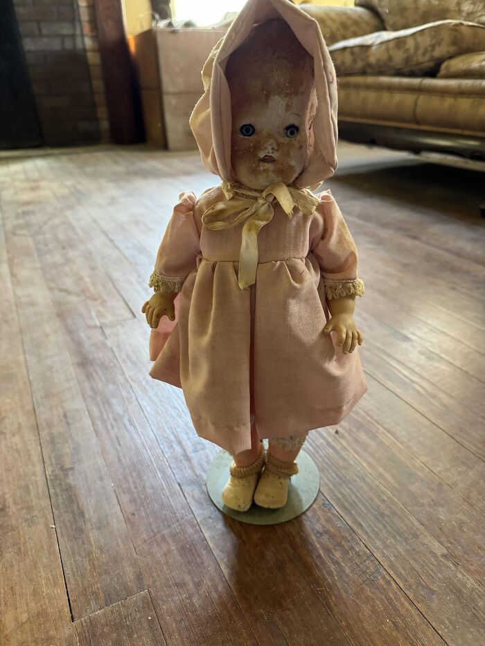 Creepy vintage doll in pink dress and bonnet found in an old home with worn wooden floors and antique furniture.