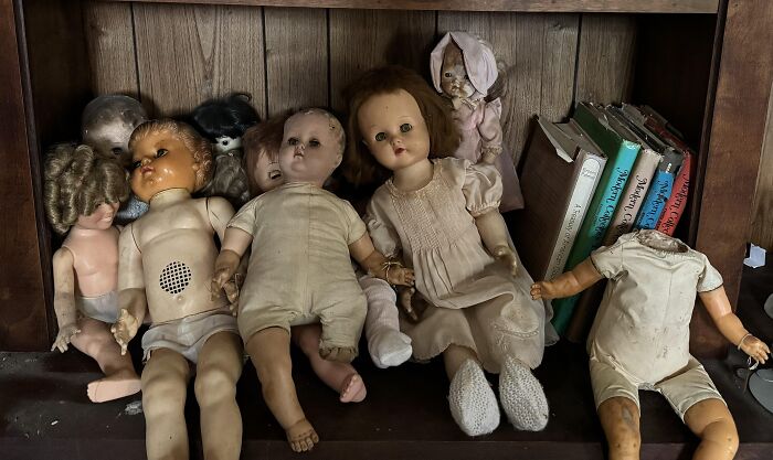 Creepy vintage dolls with worn clothes and missing parts found in an old home among dusty books on a wooden shelf.