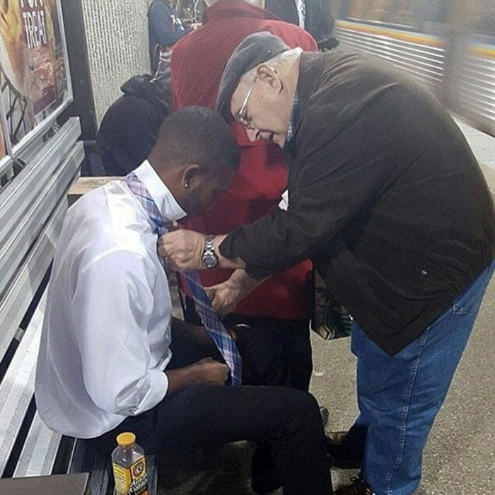 An elderly man helping a young man tie a necktie at a bus stop, showcasing uplifting acts of kindness.