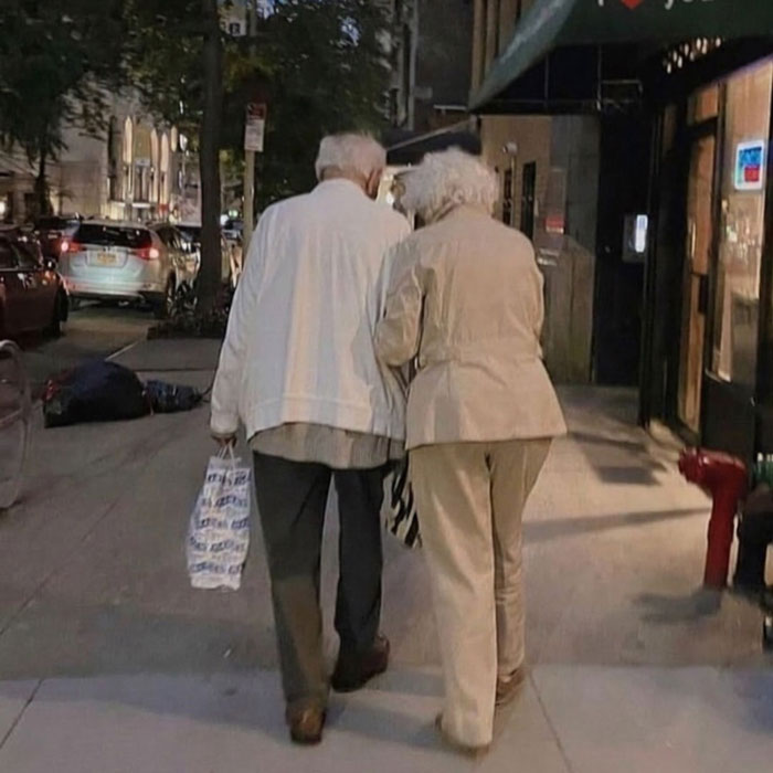 Elderly couple walking closely together on a city sidewalk at night, showing uplifting moments of kindness and love.