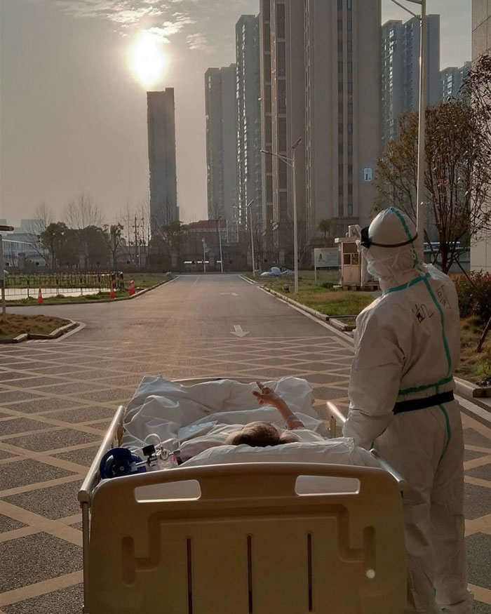 Healthcare worker in protective gear pushing patient in hospital bed under sunlight, symbolizing uplifting moments in the world.