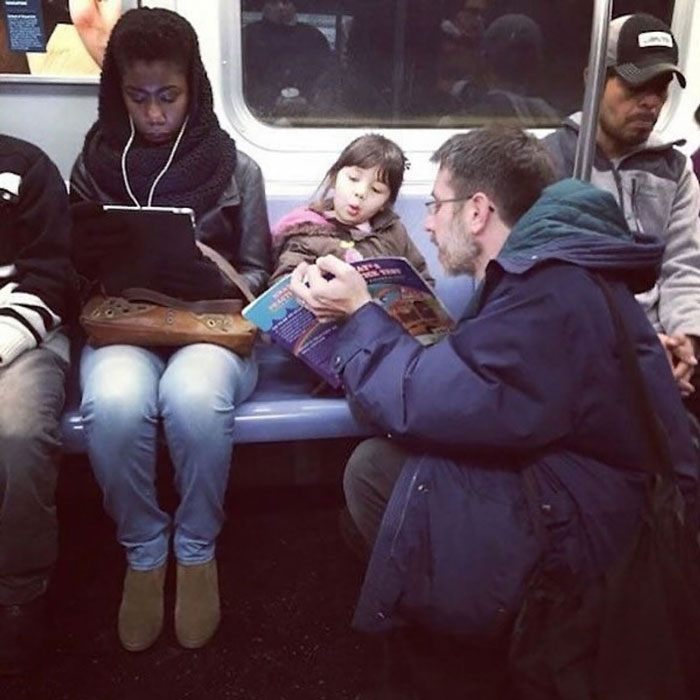 Man kneeling on subway floor reading a book to a little girl, spreading uplifting moments and kindness in the world.