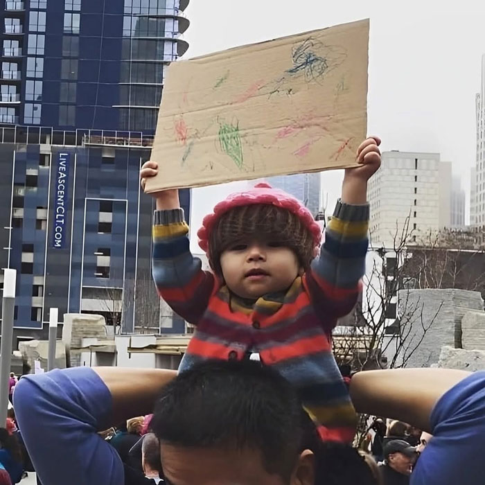 Child holding up colorful drawing on cardboard during uplifting public event in city with tall buildings behind.