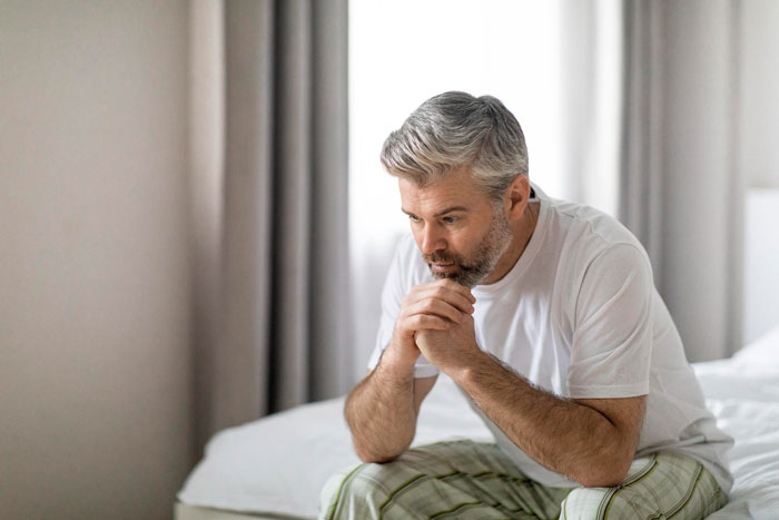 Middle-aged man sitting on bed, looking exhausted and deep in thought, reflecting on family and life challenges.