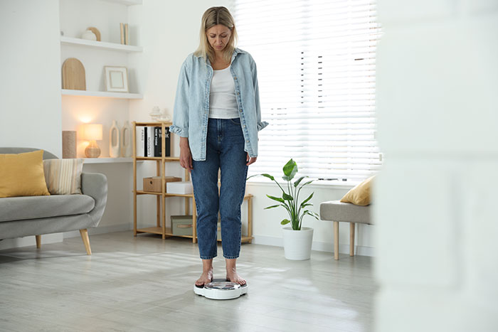 Woman looking concerned while standing on a scale in bright living room, reflecting on sister&rsquo;s weight obsession.