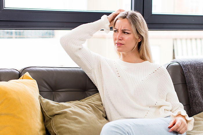 Woman sitting on couch looking confused and scratching head, depicting wife refuses to treat lice dilemma.