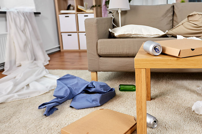 Living room mess with empty cans, pizza boxes, and clothes scattered, illustrating stress after a mom's long shift.