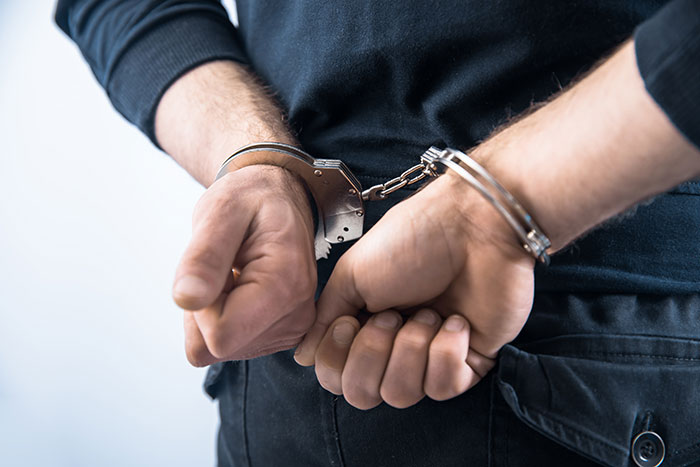 Man in handcuffs wearing a black shirt and pants with hands behind his back against a light background.