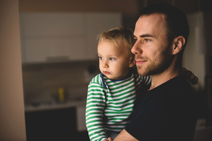 Young father holding toddler, looking thoughtfully out a window, reflecting on grandparents kicking family out over toddler.