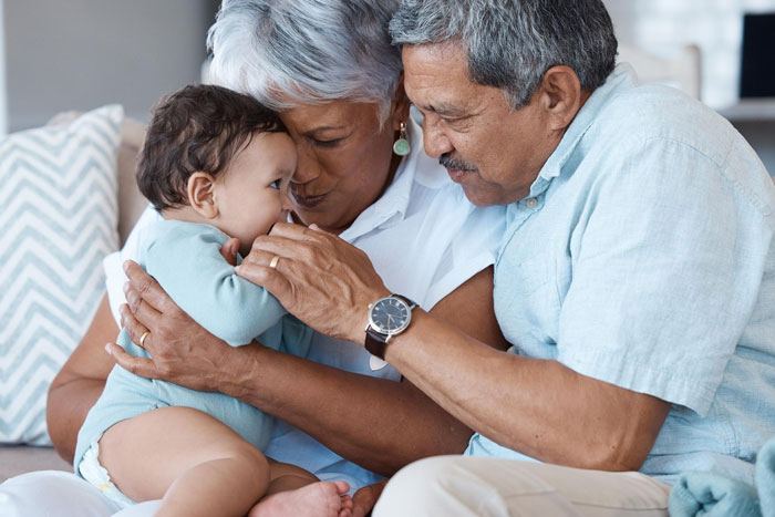 Elderly grandparents lovingly holding and playing with their toddler grandchild in a cozy home setting.