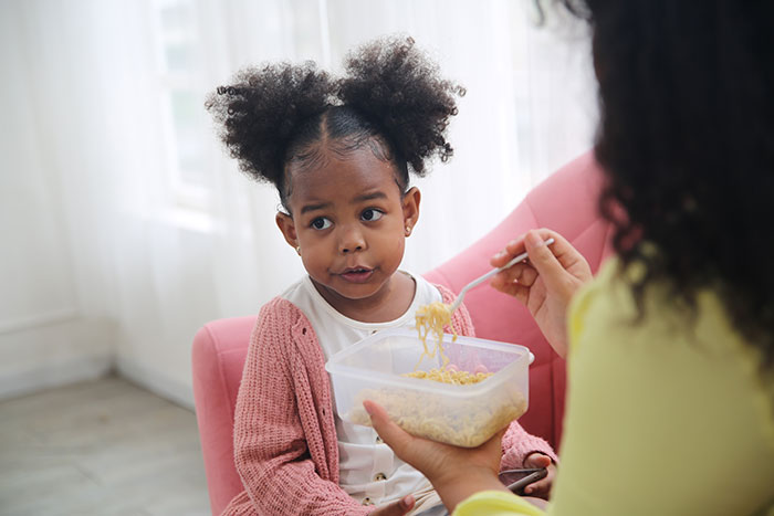 Young girl sitting unsure while woman feeds her noodles, illustrating a vegan-hating husband affecting daughter's friendship.
