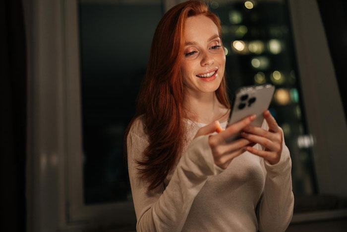Woman smiling while looking at her phone indoors at night, relating to open marriage and relationship doubts.