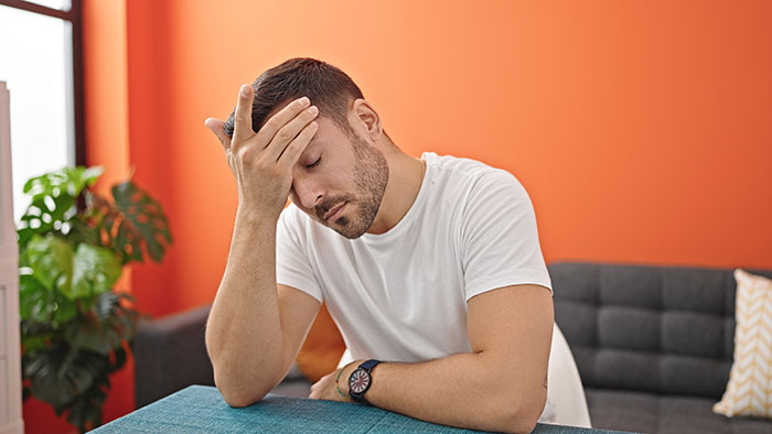 Man in white t-shirt sitting indoors with hand on forehead, looking stressed in a bright room with orange walls.