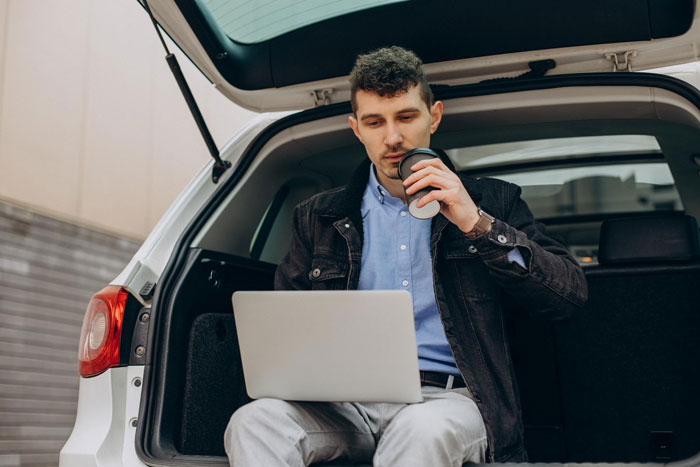 Teacher hubby working on a laptop in a car trunk, drinking coffee during a Zoom session with students.