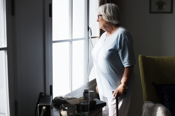 Elderly woman living alone, standing by a window with a cane, reflecting on scary encounters at home.