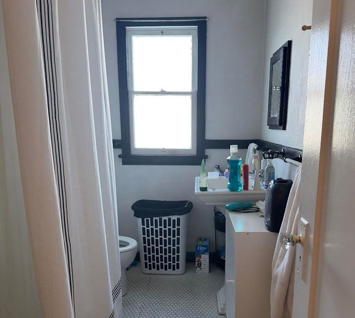 Bathroom with bright window, laundry basket, toiletries on sink, and shower curtain in a small clean space.