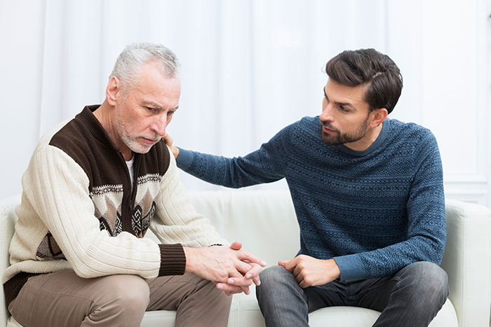 Older man looking upset while younger man consoles him on a couch, reflecting self-absorbed breakup drama at funeral.