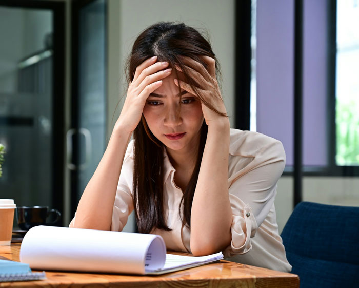 Stressed woman sitting at desk, holding head in hands, reflecting on boyfriend and pregnant mistress in front of coworkers.