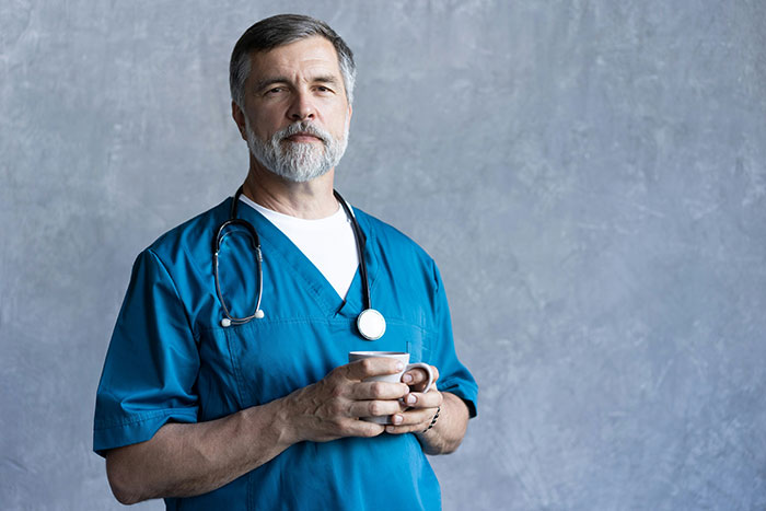 Male doctor in blue scrubs with stethoscope holding a cup, representing hospital experiences and medical care.