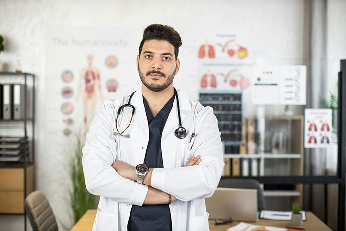 Male doctor standing with arms crossed in a hospital office, representing hospital experiences and medical care issues.