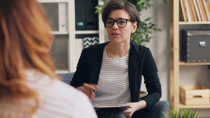 Woman in glasses and a black blazer listening attentively during a discussion about hospital experiences.