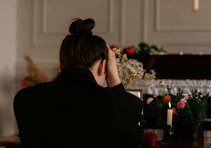Woman in black mourning attire holding her head near a candle and flowers during a memorable funeral moment.