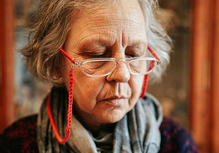 Elderly woman with glasses and gray hair looking down, reflecting on memorable funeral moments that went wrong.