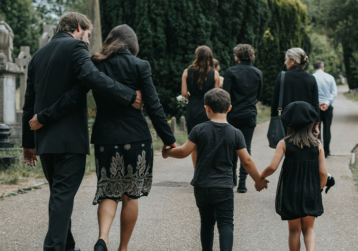 Family dressed in black walking through cemetery path, capturing memorable funeral moments with children holding hands.