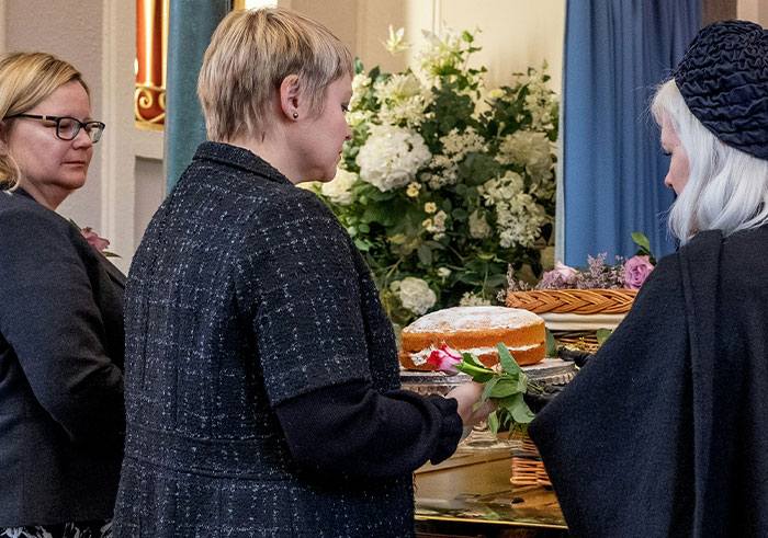 Three women at a funeral gathering, holding flowers near a table with cake and floral arrangements.