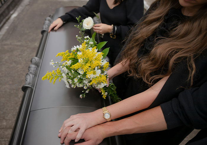 Two women in black mourning clothes holding hands and flowers next to a closed coffin during a funeral ceremony.