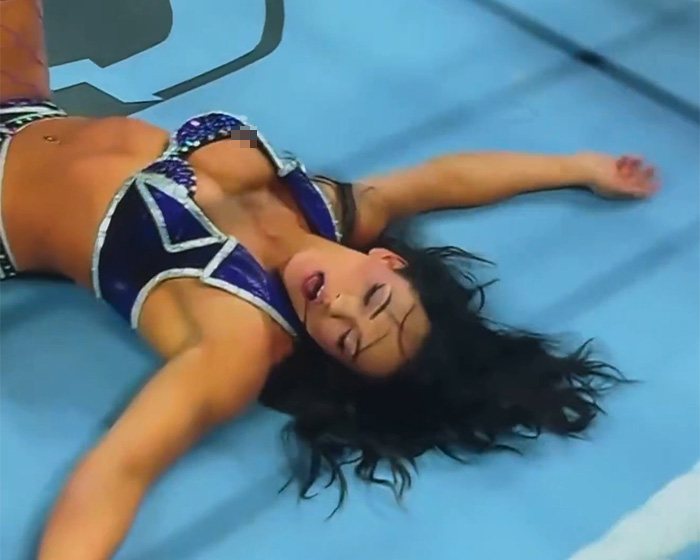 Pro wrestler Kiana James lying on the wrestling mat in a blue outfit after an awkward wardrobe malfunction on live TV.