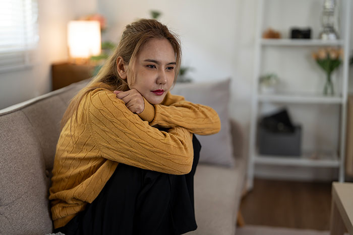 Young woman in a mustard sweater sitting on a couch looking thoughtful in a cozy living room setting.