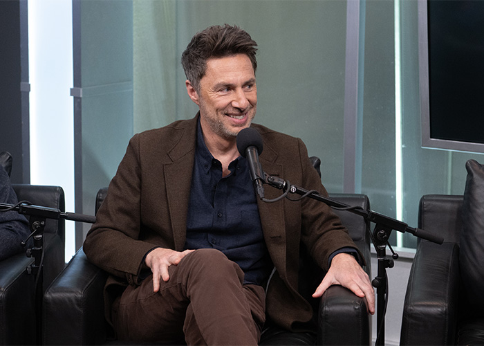 Zach Braff seated, speaking into a microphone during an interview, dressed in a brown jacket and navy shirt.