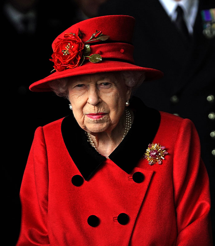 Elderly woman in a red hat and coat with a floral brooch, representing Harry and Meghan Markle turbulent dynamic.