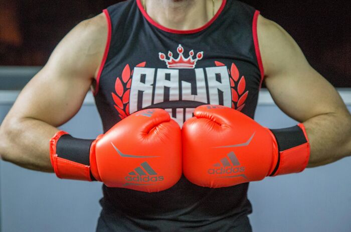 Man wearing bright red Adidas boxing gloves and a black sleeveless shirt in a gym setting, showcasing strength and focus.