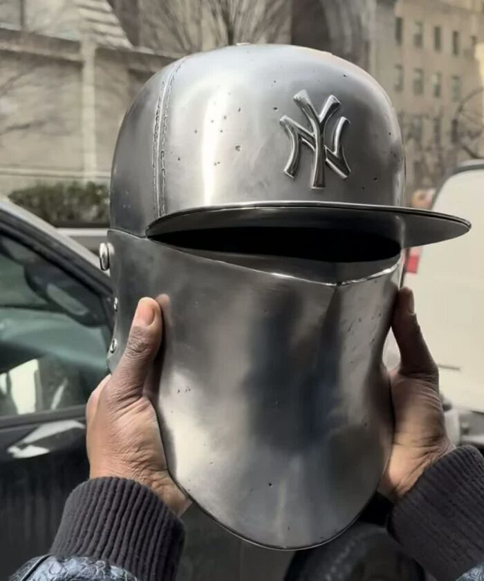 A person holding a weird metal helmet designed like a baseball cap with a NY Yankees logo. This is one of the weird images.
