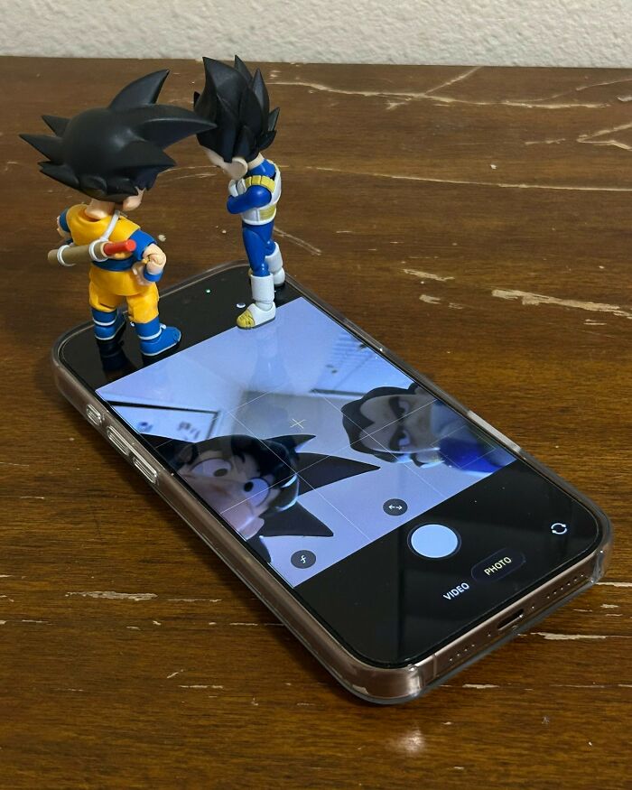 Two Dragon Ball Z figures stand on a phone screen, their reflections visible. A weird image that makes people go Hmmm.
